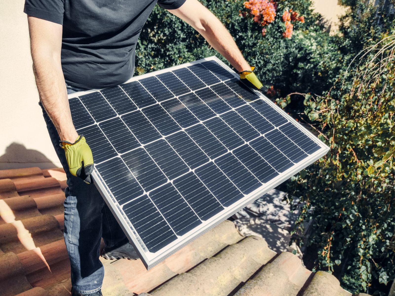 A solar installer carrying a photovoltaic panel onto a tiled residential roof under a bright sky in St. George, Utah